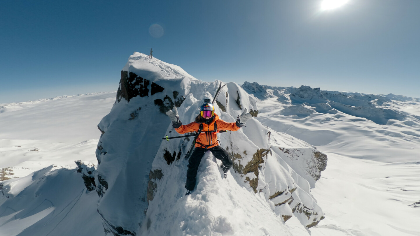 Skier in an orange jacket balancing on a narrow, snow-covered rock ledge, surrounded by a spectacular winter landscape under a bright sky.