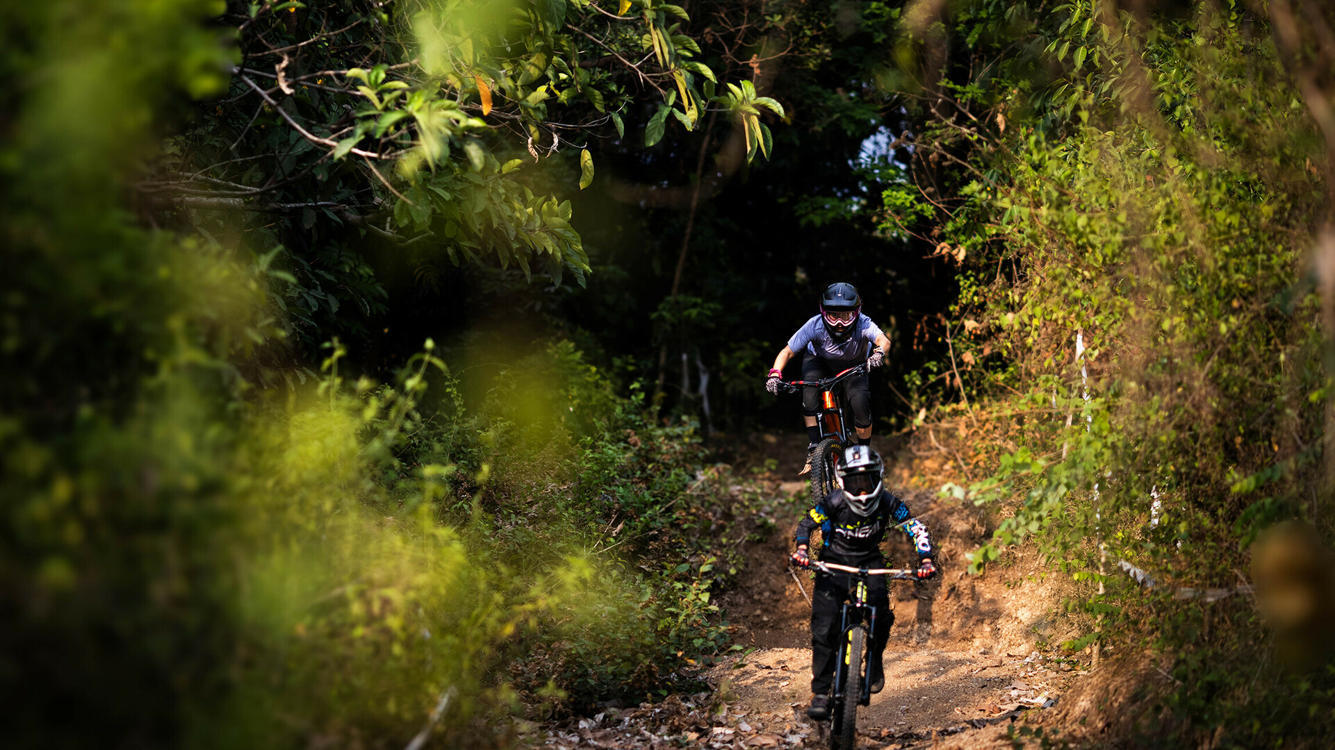 Two mountain bikers with helmets ride on a dirt road through a forest with green foliage on both sides.