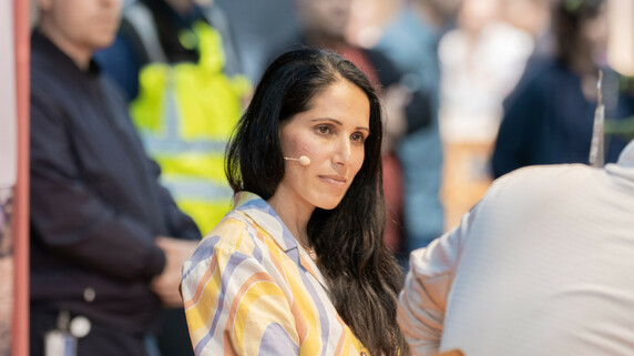 Nasim Eshqi sitting on a chair during an event, wearing an earpiece microphone. She is dressed in a colorful striped shirt. Several people are visible in the blurred background, attentively listening.